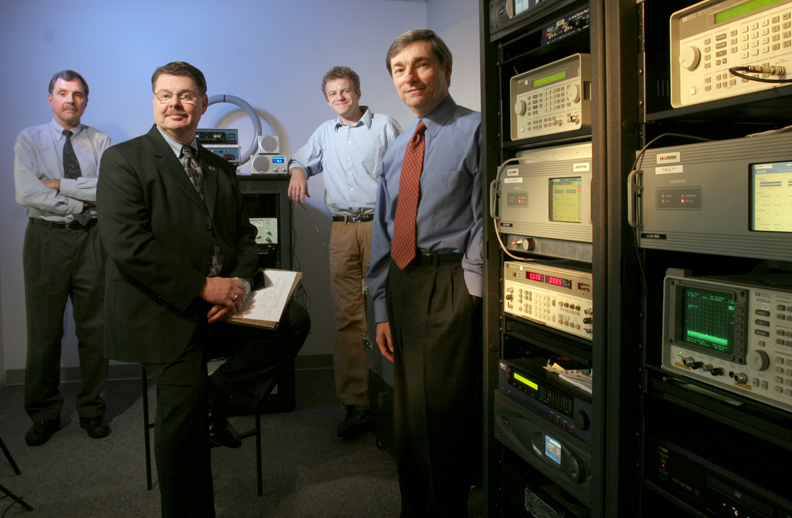 Four men standing in the new NPR lab, next to various types of electronic equipment on racks.