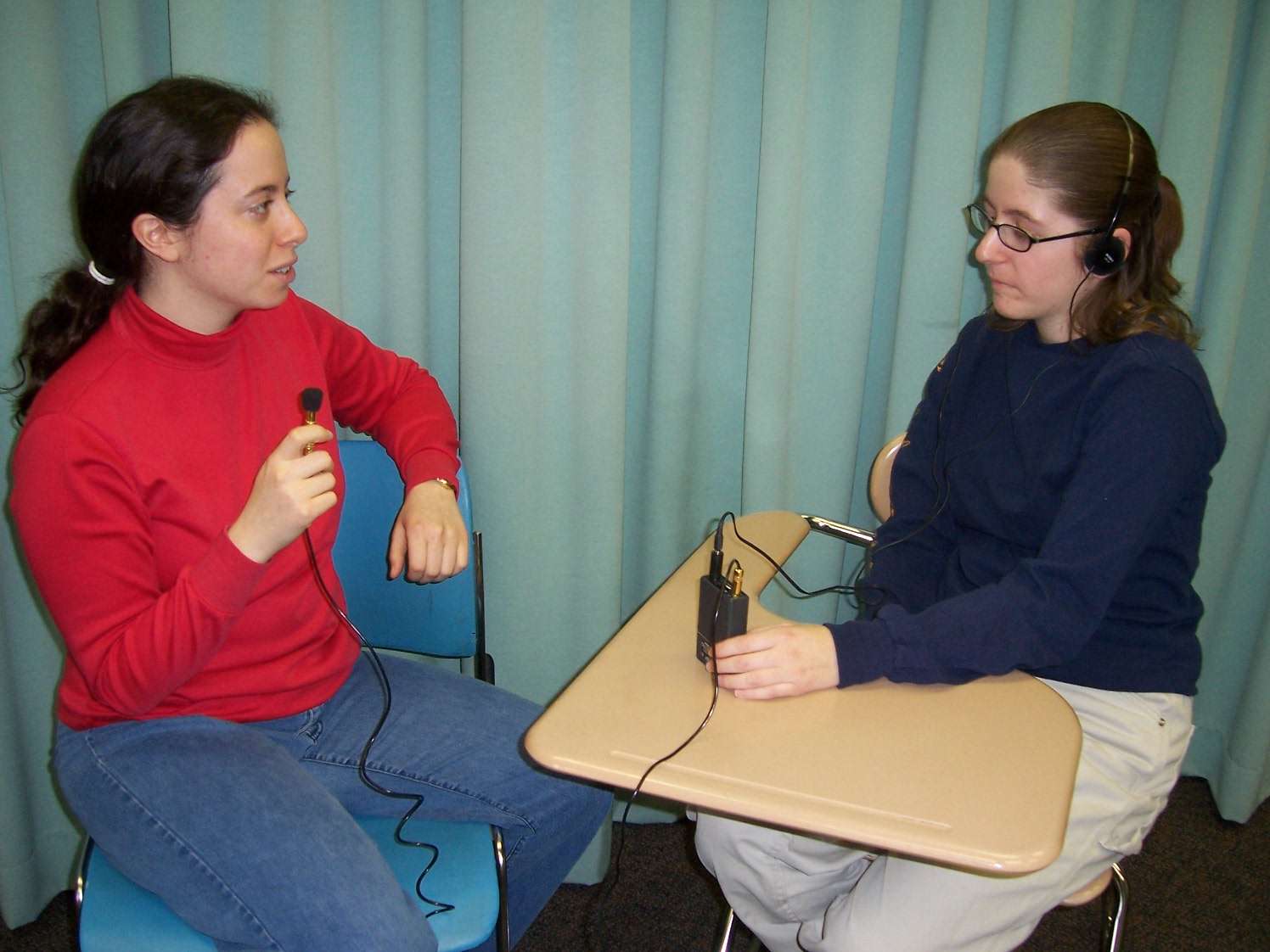 Photo of two women talking using an ALD -- one speaking into a microphone, and one wearing a headset.