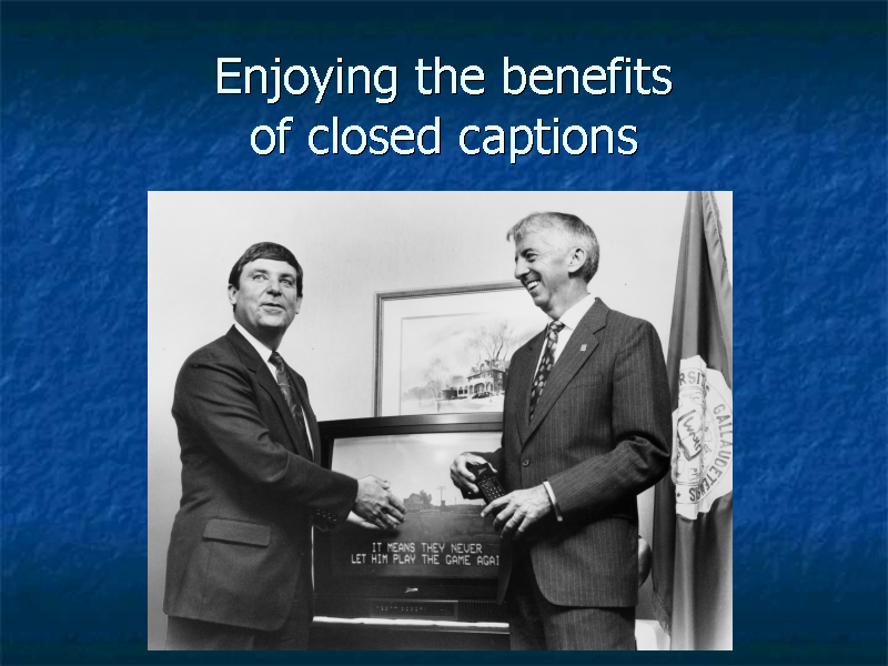 Photo of I King Jordan and another man standing in front of a  TV with captions.